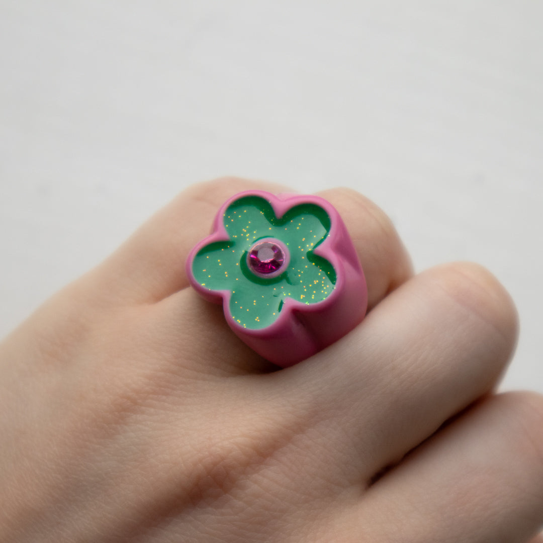 Hand wearing a pink and green flower-shaped ring with a gemstone on a white background