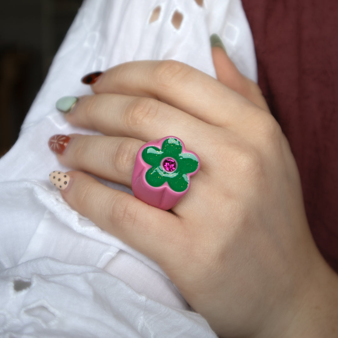 Hand wearing a pink flower-shaped ring with a green center and purple gemstone.