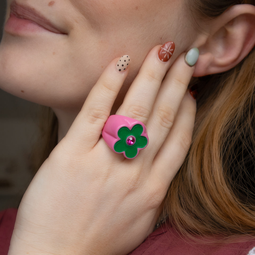 Close-up of a hand wearing a pink flower-shaped ring with a green center and red gemstone, held near an ear.